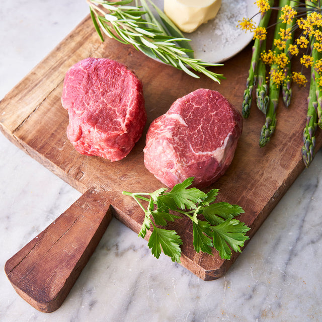 Two raw 6 oz. filet mignons sitting on a wooden cutting board next to  butter and asparagus.